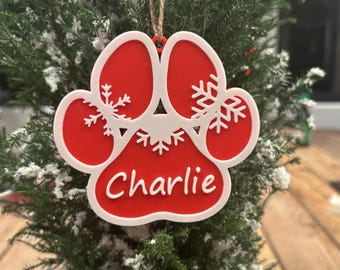 May include: A red and white paw print Christmas ornament with the name "Charlie" in white script. The ornament has snowflake cutouts and hangs from a natural twine string. The ornament is displayed on a small evergreen tree.