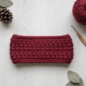 Könnte beinhalten: Ein handgefertigtes, weinrotes Häkelstirnband mit einem strukturierten Stichmuster. Ein Knäuel passender Wolle, eine Häkelnadel, ein Tannenzapfen und Eukalyptusblätter sind um das Stirnband auf einer weißen Holzoberfläche angeordnet.
