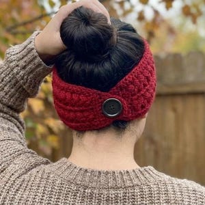 May include: A red crocheted headband with a large, dark button closure. The headband is worn by a person with dark hair pulled into a bun. The person is wearing a brown sweater.