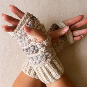 May include: A pair of cream-coloured, hand-knitted fingerless gloves. The gloves have a textured pattern with grey and brown accents. The wrist cuffs are ribbed, and the open finger design allows for dexterity. The gloves are made of a soft, warm yarn.