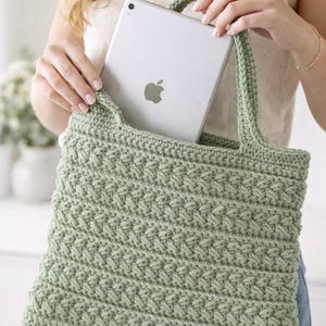 Könnte beinhalten: Eine hellgrüne gehäkelte Tragetasche mit einem strukturierten Stichmuster. Die Tasche hat zwei Griffe und hält ein silbernes Tablet mit dem Apple-Logo. Die Tasche ist handgefertigt.