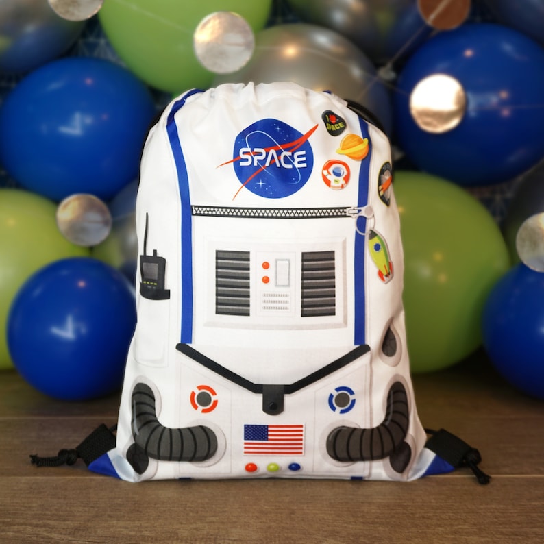 May include: White drawstring bag with a space-themed design. The bag features an astronaut with a blue and white space suit, a NASA logo, and the word "SPACE" in blue letters. The bag also has a zipper and a drawstring closure.