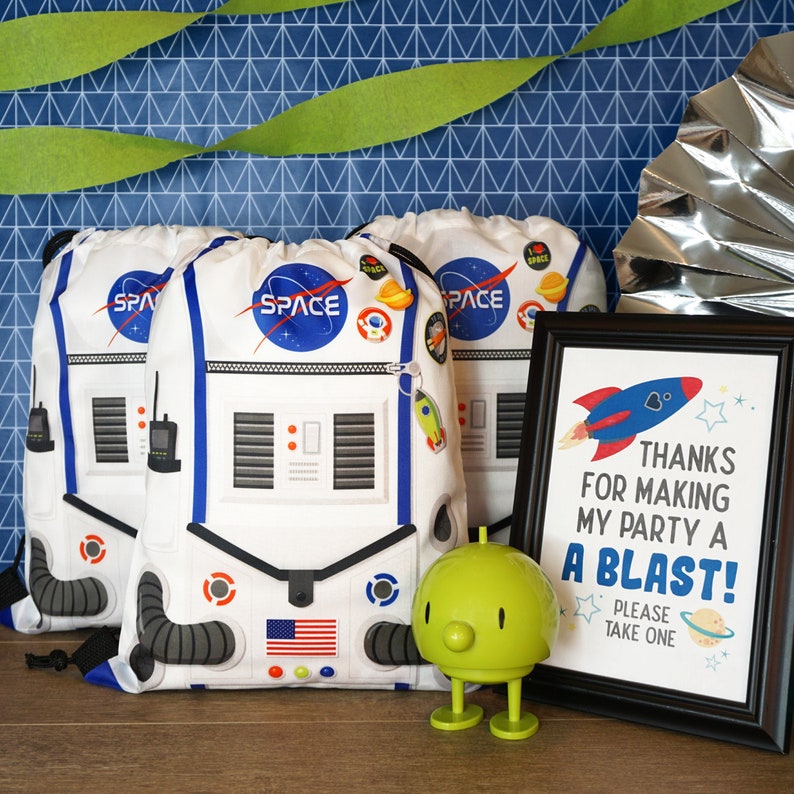 May include: Three white astronaut-themed drawstring bags with the word "SPACE" printed on them. A green alien toy and a framed sign that says "Thanks for making my party a blast! Please take one."