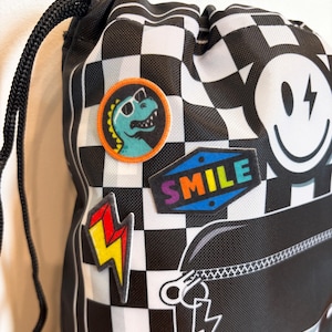 May include: A black and white checkered drawstring bag featuring a circular patch of a teal dinosaur wearing sunglasses, a diamond-shaped patch with the word SMILE in rainbow letters, a yellow and red lightning bolt patch, and a large smiley face.