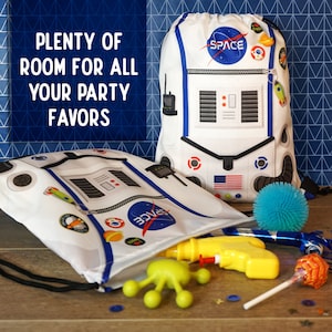 May include: Two white drawstring bags with a space-themed design. The bags have a zipper, a NASA logo, and the word "SPACE" printed on them. The bags are filled with party favors, including a water gun, a toy alien, a lollipop, and a blue spiky ball.
