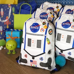 May include: Four white drawstring bags with a space theme. Each bag features a white astronaut helmet with a blue visor and the word "SPACE" in blue letters. The bags have black straps and are decorated with various space-themed patches and charms.