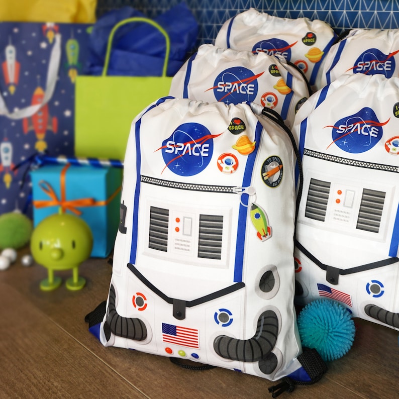 May include: Four white drawstring bags with a space theme. Each bag features a white astronaut helmet with a blue visor and the word "SPACE" in blue letters. The bags have black straps and are decorated with various space-themed patches and charms.