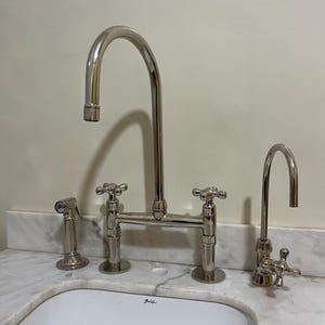 Puede incluir: Un juego de grifos de cocina cromados pulidos con un caño de arco alto, manijas en cruz y un rociador lateral. El grifo está montado sobre una encimera de mármol blanco.