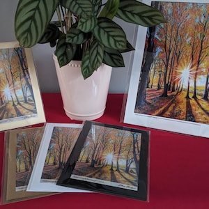 May include: Artwork prints featuring a vibrant autumn forest scene with a bright sunburst. The prints are in various sizes and frames, including a large print and smaller matted prints. A potted plant with green leaves is in the background.