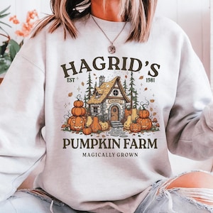 May include: A light gray crewneck sweatshirt featuring a graphic print of a stone cottage surrounded by orange pumpkins and pine trees. The text reads HAGRID'S EST 1981 PUMPKIN FARM MAGICALLY GROWN in dark brown lettering across the front of the garment.