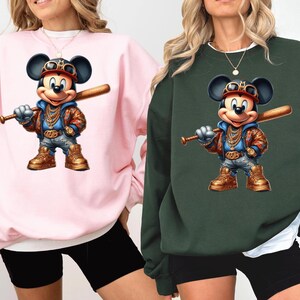 May include: Two pink and green sweatshirts with a cartoon image of Mickey Mouse wearing a gold chain and holding a baseball bat. The Mickey Mouse image is in a graffiti style.