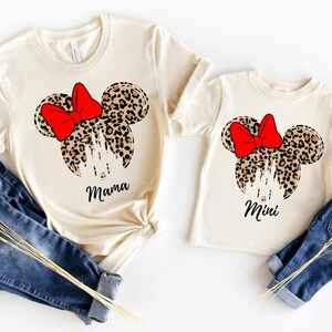 May include: Two cream-colored short-sleeve t-shirts featuring a graphic of a leopard print mouse head silhouette with a bright red bow and a white castle outline. One shirt has the word Mama printed below, and the other has the word Mini printed below.