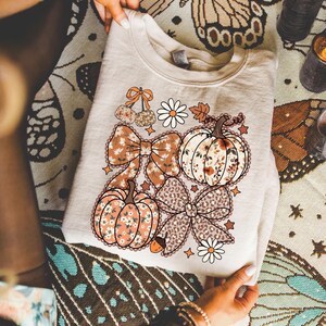 May include: A beige crewneck sweatshirt featuring a graphic print of two pumpkins, two large bows, two cherries, two daisies, and small stars. One pumpkin and one bow have a floral pattern, while the other bow has a leopard print pattern.