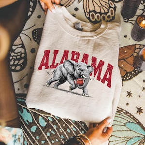 May include: A cream-colored sweatshirt featuring the word "ALABAMA" in red block letters above a gray elephant graphic holding a football. The sweatshirt is folded and held by hands.
