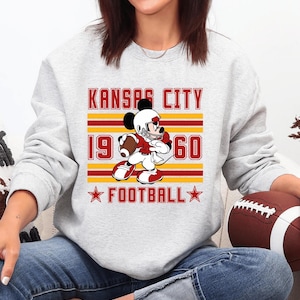 May include: A light gray sweatshirt featuring a graphic of a cartoon character in a football uniform. The graphic includes the text "KANSAS CITY 1960 FOOTBALL" in red and gold. A brown football is visible.