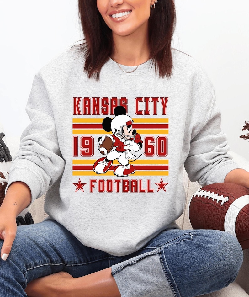 May include: A light gray sweatshirt featuring a graphic of a cartoon character in a football uniform. The graphic includes the text "KANSAS CITY 1960 FOOTBALL" in red and gold. A brown football is visible.