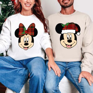 May include: Two sweatshirts featuring Mickey and Minnie Mouse. One is white with Minnie wearing a plaid bow. The other is beige with Mickey wearing a plaid Santa hat. Both have black ears and are smiling. The models are wearing blue jeans.