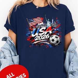 A woman wearing a navy blue t-shirt with a graphic design featuring the Statue of Liberty, an American flag, and a soccer ball, along with the text "USA 2026" and "ALL COUNTRIES".