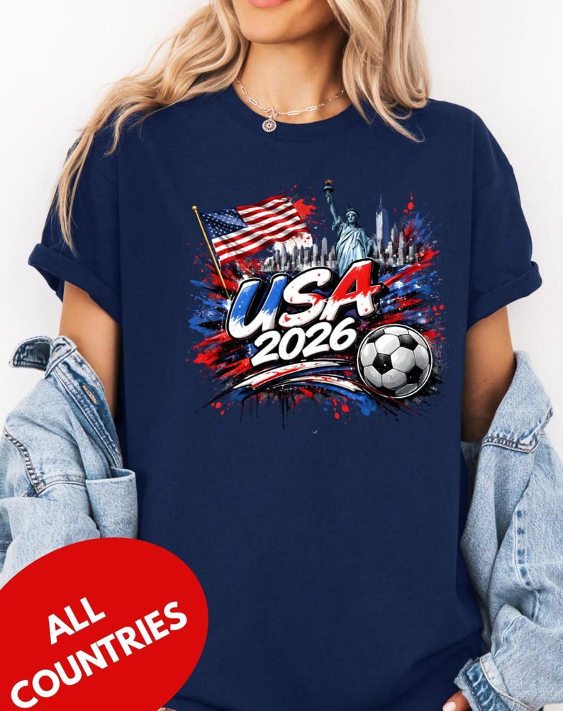 A woman wearing a navy blue t-shirt with a graphic design featuring the Statue of Liberty, an American flag, and a soccer ball, along with the text "USA 2026" and "ALL COUNTRIES".