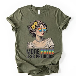 May include: An olive green t-shirt featuring a graphic of a person with curly hair adorned with yellow flowers, wearing heart-shaped rainbow sunglasses and a beaded necklace. Below the graphic, the text reads MORE PRIDE LESS PREJUDICE.