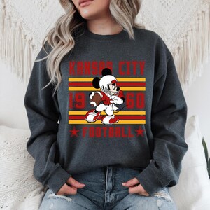 May include: Dark gray sweatshirt featuring a graphic of Mickey Mouse in a football uniform, holding a football. The design includes the text "Kansas City 1950 Football" in red and yellow. The sweatshirt has a crew neck.