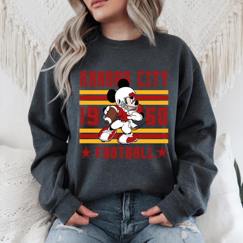 May include: Dark gray sweatshirt featuring a graphic of Mickey Mouse in a football uniform, holding a football. The design includes the text "Kansas City 1950 Football" in red and yellow. The sweatshirt has a crew neck.