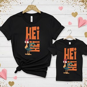 Op de afbeelding: Twee zwarte T-shirts met een oranje haan-afbeelding en het woord "Hei" erop gedrukt. Het grotere shirt heeft de haan aan de linkerkant en het woord "Hei" gestapeld aan de rechterkant. Het kleinere shirt heeft de haan aan de linkerkant en het woord "Hei" gestapeld aan de rechterkant.