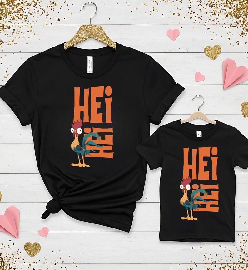 Op de afbeelding: Twee zwarte T-shirts met een oranje haan-afbeelding en het woord "Hei" erop gedrukt. Het grotere shirt heeft de haan aan de linkerkant en het woord "Hei" gestapeld aan de rechterkant. Het kleinere shirt heeft de haan aan de linkerkant en het woord "Hei" gestapeld aan de rechterkant.