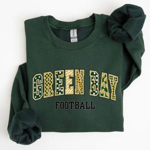 May include: A dark forest green crewneck sweatshirt featuring the text GREEN BAY in a collegiate font with varied patterns including stars, checkers, stripes, and leopard print. Below the main text, the word FOOTBALL is printed in black.