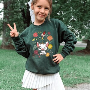 May include: A dark green sweatshirt featuring a colorful graphic of a pig, rooster, and flowers. The design is centered on the front of the sweatshirt. The sweatshirt is worn by a person.