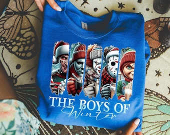 May include: A royal blue crewneck sweatshirt featuring a graphic print of five horror movie characters wearing winter hats and holding mugs. The text below the characters reads THE BOYS OF Winter in white and light blue script and block lettering.