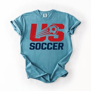 May include: A light blue t-shirt with the text "US SOCCER" in red and navy blue. The design includes a soccer ball with a trail, giving a sense of motion. The shirt is a casual wear item.