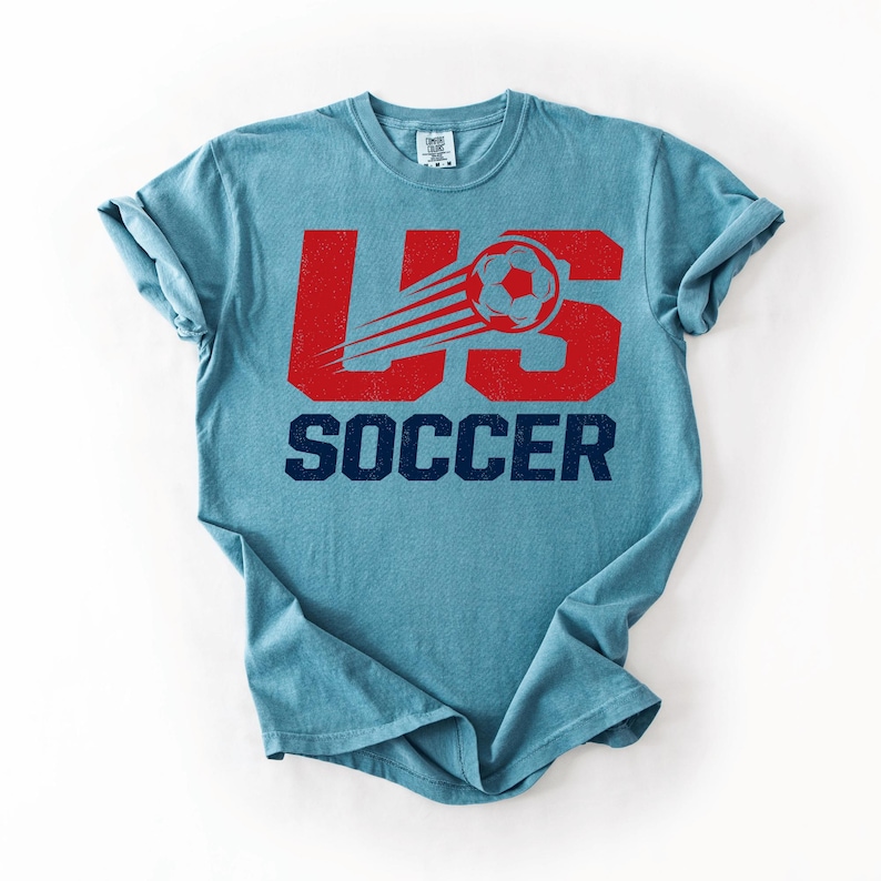May include: A light blue t-shirt with the text "US SOCCER" in red and navy blue. The design includes a soccer ball with a trail, giving a sense of motion. The shirt is a casual wear item.