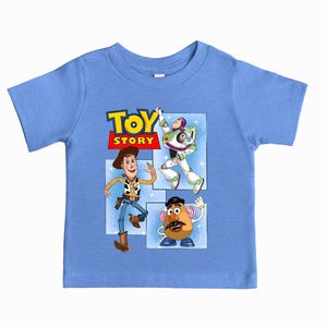May include: A light blue short-sleeve t-shirt featuring a graphic print of the Toy Story logo and characters Woody, Buzz Lightyear, and Mr. Potato Head. The design is set against a light blue background with white star accents and rectangular frames.