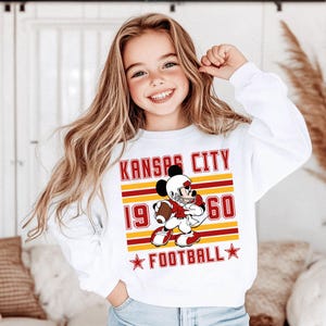 May include: White long-sleeve sweatshirt featuring a cartoon Mickey Mouse in a football uniform. The design includes the text "KANSAS CITY 1960 FOOTBALL" in red and yellow. The sweatshirt is worn by a young girl with long blonde hair.