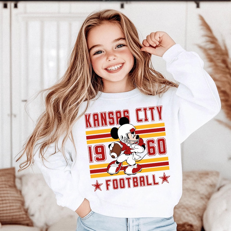 May include: White long-sleeve sweatshirt featuring a cartoon Mickey Mouse in a football uniform. The design includes the text "KANSAS CITY 1960 FOOTBALL" in red and yellow. The sweatshirt is worn by a young girl with long blonde hair.