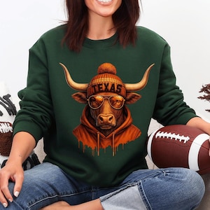 May include: A dark green sweatshirt featuring a longhorn wearing a beanie with "TEXAS" on it, sunglasses, and an orange hoodie. The longhorn is holding a football. The sweatshirt is paired with blue jeans.