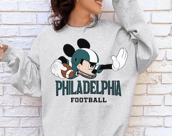May include: A light gray crewneck sweatshirt featuring a graphic of Mickey Mouse wearing a dark green and white football helmet while holding a brown football. Below the graphic, the text reads PHILADELPHIA FOOTBALL in dark green and black.