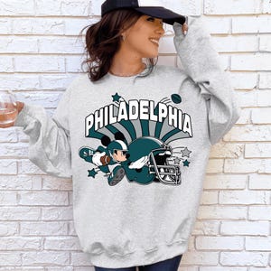May include: Gray sweatshirt with a cartoon character wearing a football helmet and holding a football. The text "PHILADELPHIA" is printed on the sweatshirt.