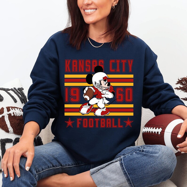 May include: Navy blue sweatshirt featuring a cartoon Mickey Mouse in a football uniform, holding a football. The text "KANSAS CITY 1960 FOOTBALL" is printed in red and yellow. A football is held in the person's hands.