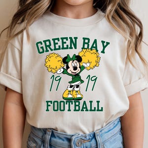 May include: A beige t-shirt featuring a cartoon Minnie Mouse dressed in a green cheerleader outfit, holding yellow pom-poms. The text "GREEN BAY FOOTBALL" and the year "1919" are printed above and below the character in green.