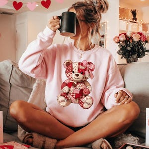 May include: A light pink sweatshirt featuring a patchwork teddy bear design with a heart that says "Love." The bear has a pink bow and the words "Hugs" and "Kisses" on its feet. A black mug is held up to the face.