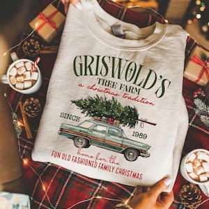 May include: A light gray sweatshirt with the text "Griswold's Tree Farm, a Christmas tradition" and a graphic of a car carrying a Christmas tree. The sweatshirt also has the text "Since 1989" and "Home for the fun old fashioned family Christmas".