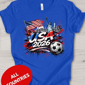A blue t-shirt with a graphic design featuring the Statue of Liberty, an American flag, and a soccer ball, along with the text "USA 2026" and a red circle with the text "ALL COUNTRIES" below the design.