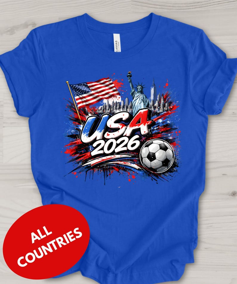 A blue t-shirt with a graphic design featuring the Statue of Liberty, an American flag, and a soccer ball, along with the text "USA 2026" and a red circle with the text "ALL COUNTRIES" below the design.
