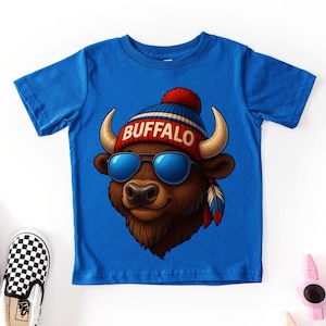 May include: A bright blue short-sleeved t-shirt featuring a graphic of a brown buffalo wearing blue sunglasses and a red, white, and blue knit cap with a pom-pom. The word BUFFALO is printed in white letters on the front of the cap.