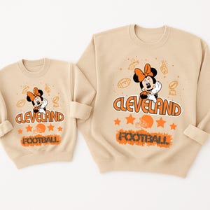 May include: Two beige sweatshirts with a cartoon Minnie Mouse design. The design includes the text "Cleveland Football", footballs, stars, and a football helmet. The sweatshirts have a crew neck and long sleeves.