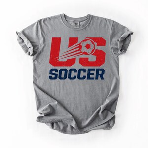 May include: Gray t-shirt with a distressed red "US" graphic and a soccer ball with speed lines. The word "SOCCER" is printed in navy blue below the graphic. The shirt is made of a soft material.