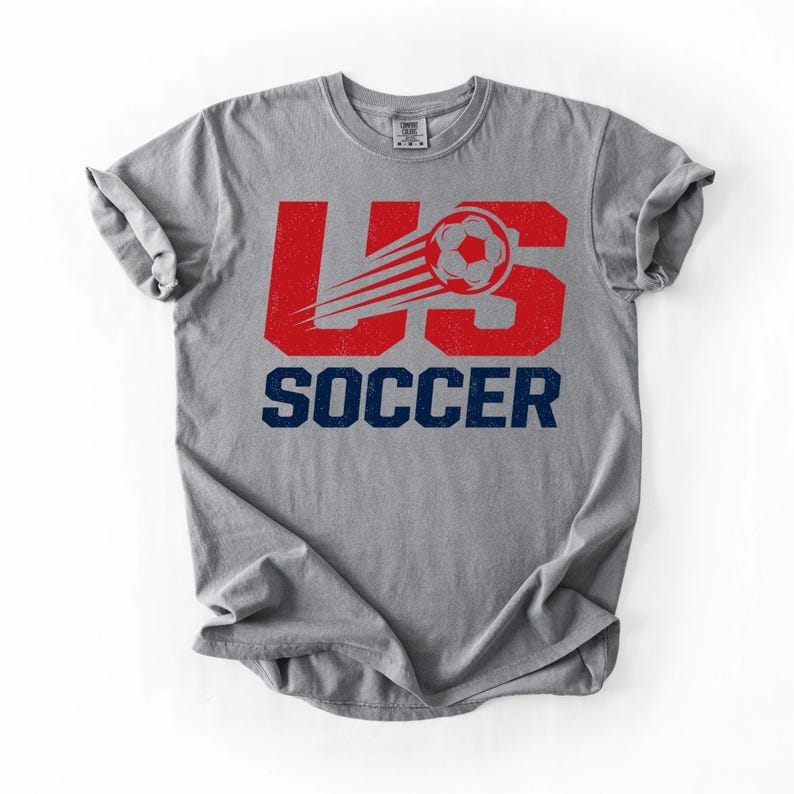 May include: Gray t-shirt with a distressed red "US" graphic and a soccer ball with speed lines. The word "SOCCER" is printed in navy blue below the graphic. The shirt is made of a soft material.
