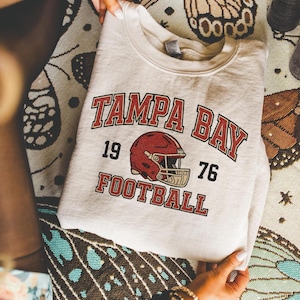 May include: A beige crewneck sweatshirt featuring a red and black graphic print that reads TAMPA BAY FOOTBALL with the year 1976 and an illustration of a red football helmet centered between the numbers. The text is in a bold, collegiate font.
